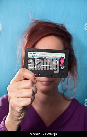 Eine Frau, die ein altes AGFA CRII Audio-Kassettenband in der Nähe ihres Gesichtes hält. Das Vintage-Band vermittelt ein Gefühl von Nostalgie und Retro-Vibes und hebt Erinnerungen aus der Vergangenheit hervor. Stockfoto