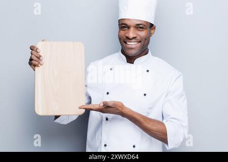 Sonderangebot vom Koch. Selbstbewusster, junger afrikanischer Koch in weißer Uniform, der ein hölzernes Schneidebrett hält und mit einem Lächeln darauf zeigt, während er gegen ihn steht Stockfoto