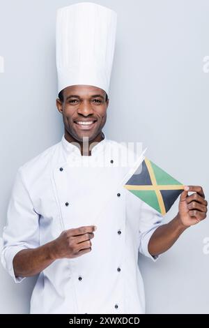 Liebe es, nationale Gerichte zu kochen. Selbstbewusster junger afrikanischer Koch in weißer Uniform, der die Flagge Jamaikas hält und vor grauem Hintergrund lächelt Stockfoto