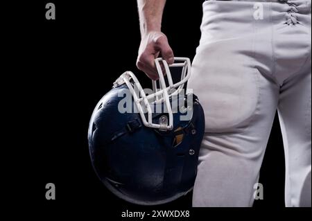 Crashhelm. Zugeschnittenes Bild eines Fußballspielers, der einen Fußballhelm hält, während er vor schwarzem Hintergrund steht Stockfoto