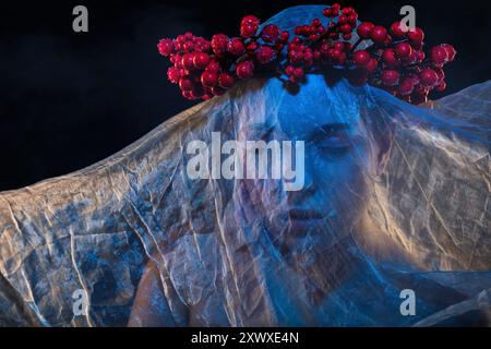 Ein Mädchen mit wunderschönem Make-up, ihr Gesicht ist bedeckt mit blauem Tüll, ein Kranz voller Beeren auf dem Kopf. Halloween-Konzept. Stockfoto