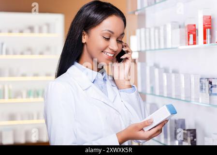 Beratung für ein geeignetes Medikament. Schöne junge Afrikanerin im Labormantel, die Behälter mit etwas Arznei hält und auf dem Handy telefoniert, während sie steht Stockfoto