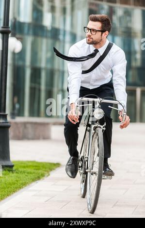 Schnell ins Büro. Voller Länge eines hübschen jungen Geschäftsmannes, der wegschaut, während er auf seinem Fahrrad fährt Stockfoto