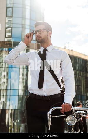Vertrauen in die Innenstadt. Niedriger Blickwinkel eines selbstbewussten jungen Geschäftsmannes, der im Freien seine Brille anpasst und die Hand am Fahrrad hält Stockfoto