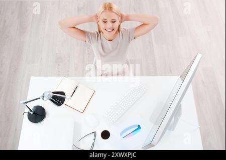 Machen Sie eine Minute Pause. Blick von oben auf die schöne junge lächelnde Frau, die Hände hinter dem Kopf hält, während sie an ihrem Arbeitsplatz sitzt Stockfoto