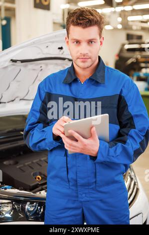 Konzentrierte sich auf Ihr Problem. Selbstbewusster junger Mann, der an einem digitalen Tablet arbeitet und die Kamera ansieht, während er in der Werkstatt steht und ein Auto im Hintergrund hat Stockfoto