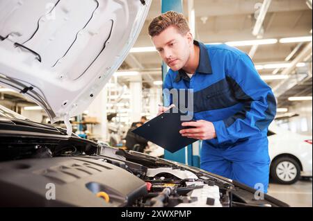 Konzentriert sich auf die Arbeit. Konzentrierter junger Mann in Uniform, der Auto untersucht und etwas in die Zwischenablage schreibt, während er in der Werkstatt steht Stockfoto