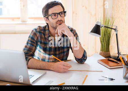 Warten auf Inspiration. Ein nachdenklicher junger Mann, der die Hand am Kinn hält und wegblickt, während er an seinem Arbeitsplatz sitzt Stockfoto