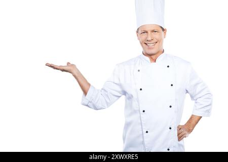 Kopieren Sie Platz in seiner Hand. Der selbstbewusste, reife Koch in weißer Uniform hält den Kopierraum in der Hand und lächelt, während er vor weißem Hintergrund steht Stockfoto