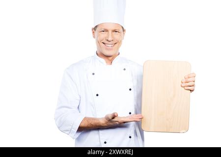 Sonderangebot vom Koch. Selbstbewusster, reifer Koch in weißer Uniform, der ein hölzernes Schneidebrett hält und mit einem Lächeln darauf zeigt, während er gegen weiß steht Stockfoto