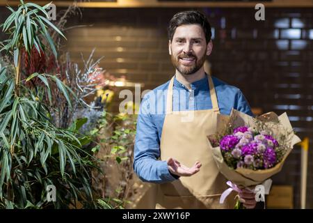 Der lächelnde Blumenhändler auf Schürze hält einen lebhaften Blumenstrauß im Blumenladen, umgeben von üppigen Pflanzen. Das Bild fängt das Wesen des Blumengeschäfts, der Kreativität und der Leidenschaft für Blumen in einer warmen, einladenden Umgebung ein. Stockfoto