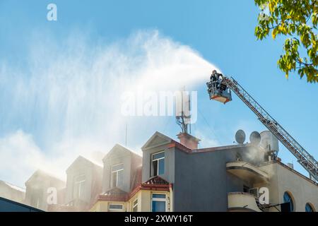 Die Feuerwehr löscht das brennende Haus mithilfe einer verlängerten Leiter, die mit einem Wasserschlauch ausgestattet ist, um Wasser mit Rauch auf das Gebäudedach zu sprühen. Feuerwehrmann Stockfoto