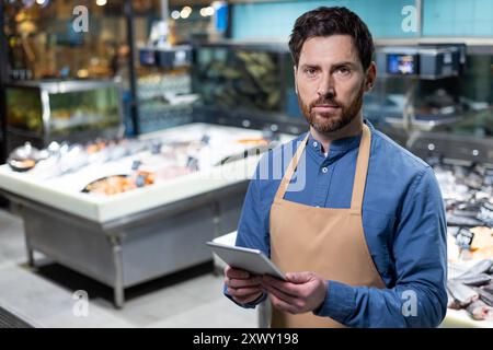 Fischmarktarbeiter in Schürze, der Tablette in der Hand hält. Stehen Sie selbstbewusst vor frischen Meeresfrüchten mit verschiedenen Fisch- und Meeresfrüchten. Repräsentiert Rollen im Einzelhandel und in der Lebensmittelindustrie. Stockfoto