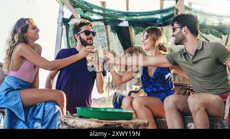 Eine Gruppe junger Freunde genießt Drinks und toast bei einer Sommerparty am Strand. Fröhliche Menschen, die sich draußen unterhalten, Leben und Freundschaft feiern Stockfoto