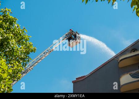 Feuerwehrleute auf einer erhöhten Plattform löschen brennendes Haus mit einer verlängerten Leiter, die mit einem Wasserschlauch ausgestattet ist, um Wasser auf das Gebäudedach zu sprühen Stockfoto