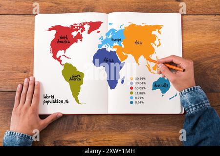 Infografik zur Weltbevölkerung. Draufsicht Nahaufnahme eines Mannes, der Hände auf seinem Notizbuch hält, mit farbenfroher Karte darauf, während er an den hölzernen des sitzt Stockfoto