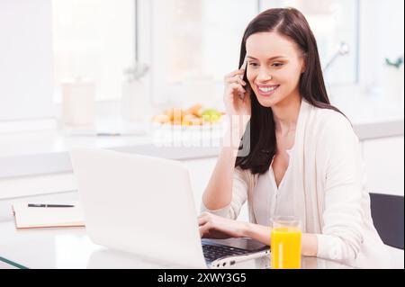 Die Möglichkeit haben, überall zu arbeiten. Attraktive junge Frau, die am Laptop arbeitet und mit dem Handy spricht, während sie in der Küche sitzt Stockfoto