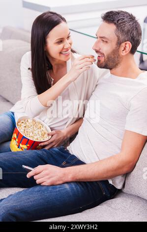 Genießen Sie unbeschwerte gemeinsame Zeit. Wunderschönes junges, liebevolles Paar, das sich aneinander knüpft und Popcorn isst, während man auf der Couch sitzt und fernsieht Stockfoto