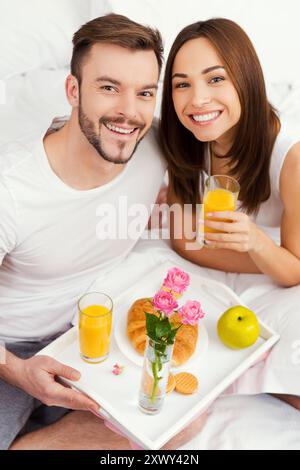 Beginnen Sie einen guten Tag mit Frühstück im Bett. Blick von oben auf ein wunderschönes junges, liebevolles Paar, das im Bett frühstückt und lächelt Stockfoto