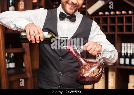 Wein in den Dekanter gießen. Zugeschnittenes Bild eines selbstbewussten männlichen Sommeliers, der Rotwein in die Karaffe gießt, während er in der Nähe des Weinregals steht Stockfoto