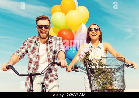 Das macht so viel Spaß! Niedriger Blickwinkel des fröhlichen jungen Paares, das lächelnd und auf Fahrrädern mit bunten Ballons im Hintergrund fährt Stockfoto