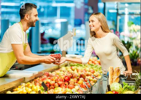 Los geht's! Seitenansicht eines jungen Kassierers, der weiblichen Kunden Himbeeren schenkt, während er im Lebensmittelgeschäft steht Stockfoto