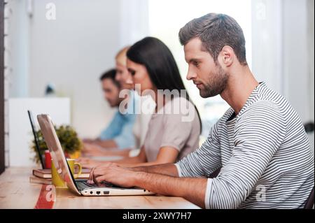 Konzentriert sich auf die Arbeit. Eine Gruppe selbstbewusster Geschäftsleute in eleganter Freizeitkleidung, die an ihren Laptops arbeiten, während sie am Arbeitsplatz in einer Reihe sitzen Stockfoto