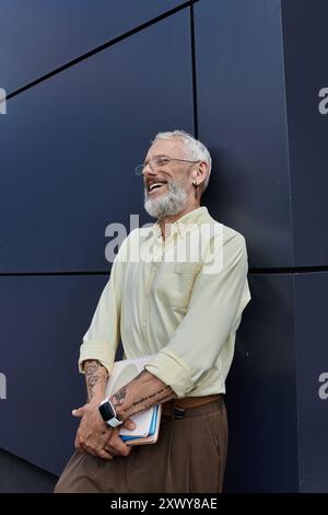 Ein Mann mittleren Alters mit Bart lacht, während er sich an ein Gebäude lehnt und einen Stapel Papiere hält. Stockfoto