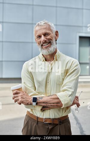 Ein reifer Mann mit Bart und warmem Lächeln steht draußen und hält eine Kaffeetasse in der Hand. Er trägt ein hellgelbes Hemd und eine braune Hose. Stockfoto