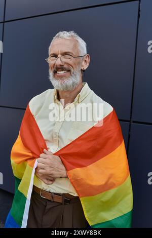 Ein reifer Schwuler, lächelnd und in eine Regenbogenfahne gehüllt, steht selbstbewusst vor einem modernen Gebäude. Stockfoto