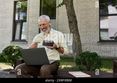 Ein reifer schwuler Mann mit Bart sitzt auf einer Bank vor einem modernen Gebäude und lacht, während er sein Handy und seinen Laptop benutzt. Stockfoto