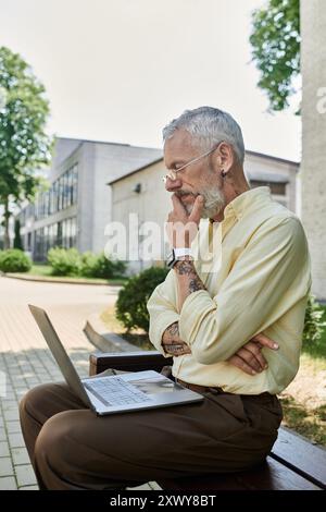 Ein reifer Mann mit Bart sitzt auf einer Bank vor einem modernen Gebäude, mit seinem Laptop und tief in Gedanken. Stockfoto