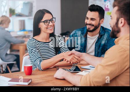 Willkommen an Bord! Selbstbewusste junge Frau und Mann, die Hände schütteln und lächeln, während sie am Schreibtisch im Büro sitzen Stockfoto