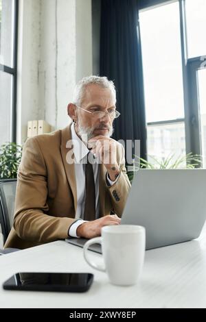 Ein lgbtq-Mann mittleren Alters mit Bart, Brille und Anzugsjacke, sitzt an einem Schreibtisch vor einem Laptop und denkt darüber nach Stockfoto