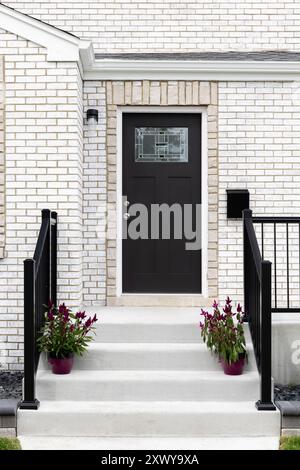 Eine Eingangstür in einem weißen Backsteinhaus mit Betontreppen und schwarzem Geländer führt zu einer schwarzen Eingangstür mit Mosaikfenster. Stockfoto