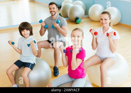 Genießen Sie Zeit in einer Art Club. Blick von oben auf eine glückliche, sportliche Familie, die mit Kurzhanteln im Sportclub trainiert, während sie zusammen auf den Fitnessbälle sitzt Stockfoto