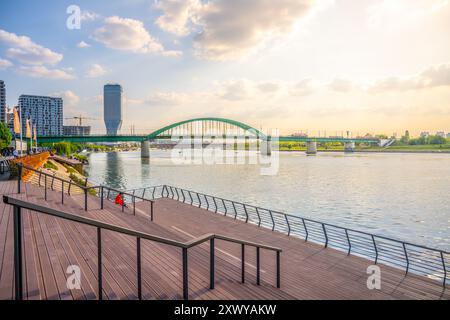 Die alte Save-Brücke überspannt die Save in Belgrad mit ihren grünen Bögen. Eine hölzerne Promenade verläuft entlang des Flusses, die die ruhige Atmosphäre am Wasser während des Sonnenuntergangs unterstreicht. Stockfoto