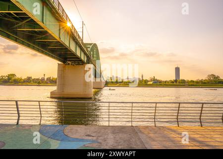 Die Sonne untergeht hinter der Branko-Brücke und wirft goldenes Licht über den Fluss Save. Das ruhige Wasser spiegelt die leuchtenden Farben des Himmels wider, während die Menschen einen Spaziergang am Fluss entlang der Promenade genießen. Stockfoto