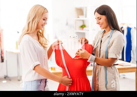 Sie achten auf Details. Zwei lächelnde junge Frauen, die in ihrer Modewerkstatt ihre Kleider messen und anpassen Stockfoto