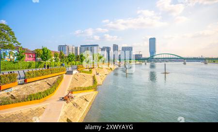 Besucher genießen einen sonnigen Tag entlang der Sava Promenade, mit modernen Gebäuden im Hintergrund und einer lebhaften Atmosphäre in der Nähe des Ufers. Stockfoto