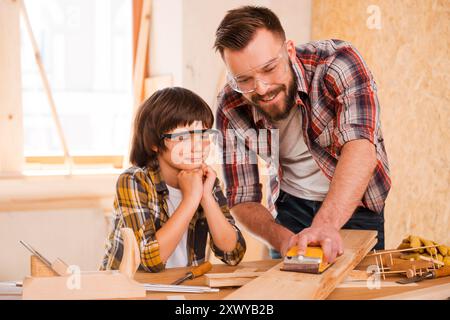 Präzision und Perfektion. Ein lächelnder junger Zimmermann zeigt seinem Sohn, wie man Holz in seiner Werkstatt anschleift Stockfoto