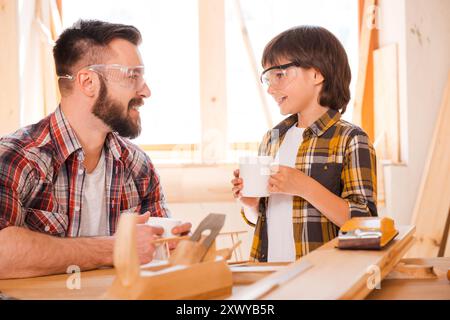 Es ist Zeit für eine Pause. Ein lächelnder junger Zimmermann und sein Sohn halten sich Tassen und schauen sich gegenseitig an, während sie in der Werkstatt arbeiten Stockfoto