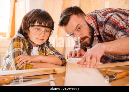 Präzision ist der wichtigste Teil der Tischlerei. Konzentrierter junger Zimmermann, der seinem Sohn in der Werkstatt zeigt, wie man mit Holz arbeitet Stockfoto