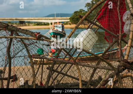 Fischfallen stapelten sich am Ufer mit einem Fischerboot im Hintergrund. Leere Fallen, die sich am Meer auftürmen und zum Angeln bereit sind. Stockfoto