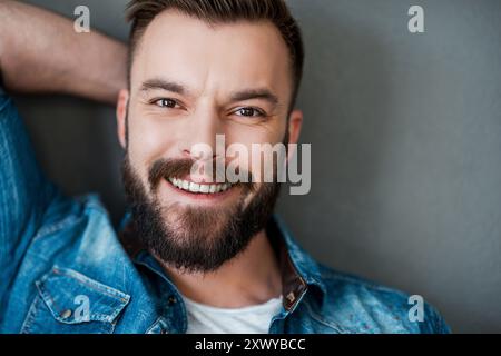 Glück kommt nie aus der Mode. Fröhlicher junger Mann, der Hand hinter Kopf hält und in die Kamera lächelt, während er vor grauem Hintergrund steht Stockfoto