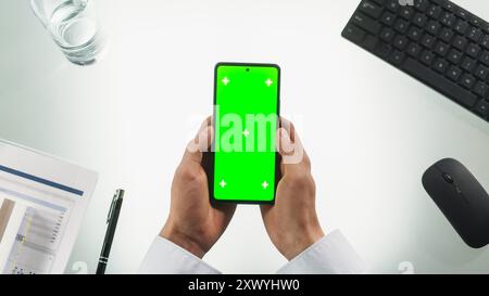 Anonyme Person, die ein Smartphone mit Mock-up-Chromakey-Display mit grünem Bildschirm und Platzhaltern verwendet. Mann hält das Mobiltelefon mit beiden Händen vertikal, wischt und tippt auf das Telefon. Top-Down-Ansicht Stockfoto