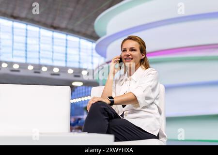 Selbstbewusste junge Geschäftsfrau sitzt in der Halle des Geschäftszentrums und spricht mit ihrem Handy. Stockfoto