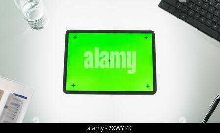 Draufsicht eines Tablet-Computers mit Mock-up-grünem Chromakey-Display mit Motion Tracker auf einem Schreibtisch neben Tastatur, schwarzem Stift, Dokumenten und einem Glas Wasser Stockfoto