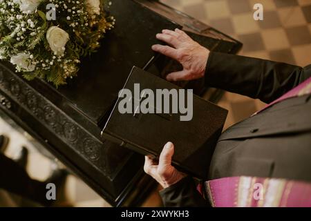 Hochwinkelaufnahme eines älteren, nicht erkennbaren Pastors in schwarzem Gewand, der schwarze Bibel hält, mit einem Kreuz auf der Abdeckung, das schwarzen Holzsarg berührt Stockfoto
