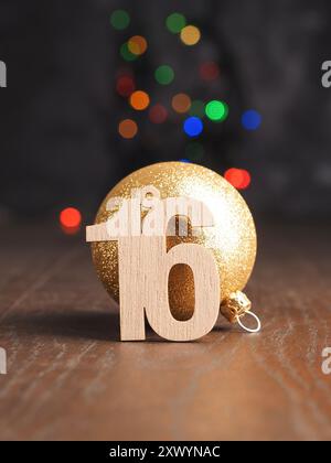 16. Dezember, hölzerne Nummer 16 mit einer goldenen Weihnachtskugel auf verschwommenen Weihnachtslichtern Stockfoto